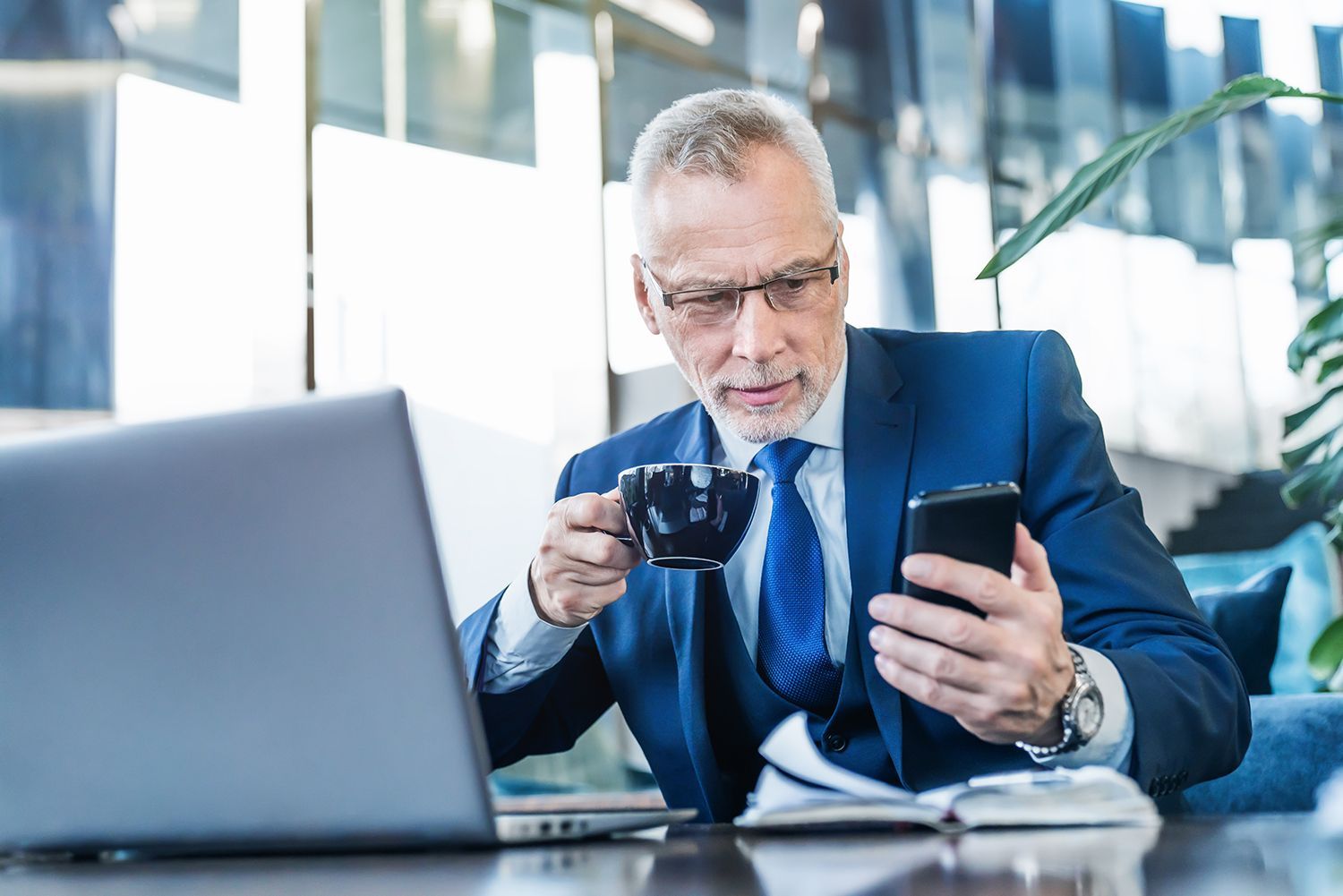 A man in a suit is sitting at a table drinking coffee and looking at his cell phone.