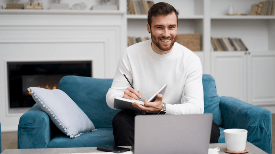 A man is sitting on a couch with a laptop and a notebook.