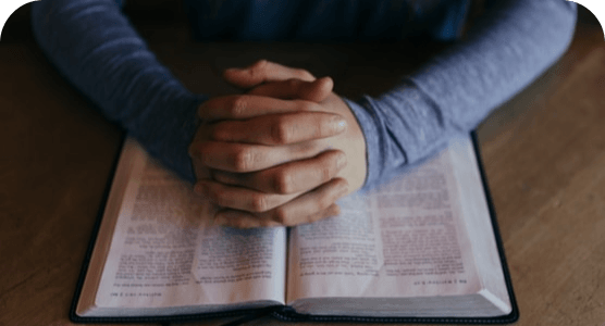 A person is sitting at a table with their hands folded over an open bible.