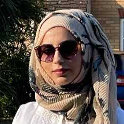A woman wearing a hijab and sunglasses is standing in front of a brick building.