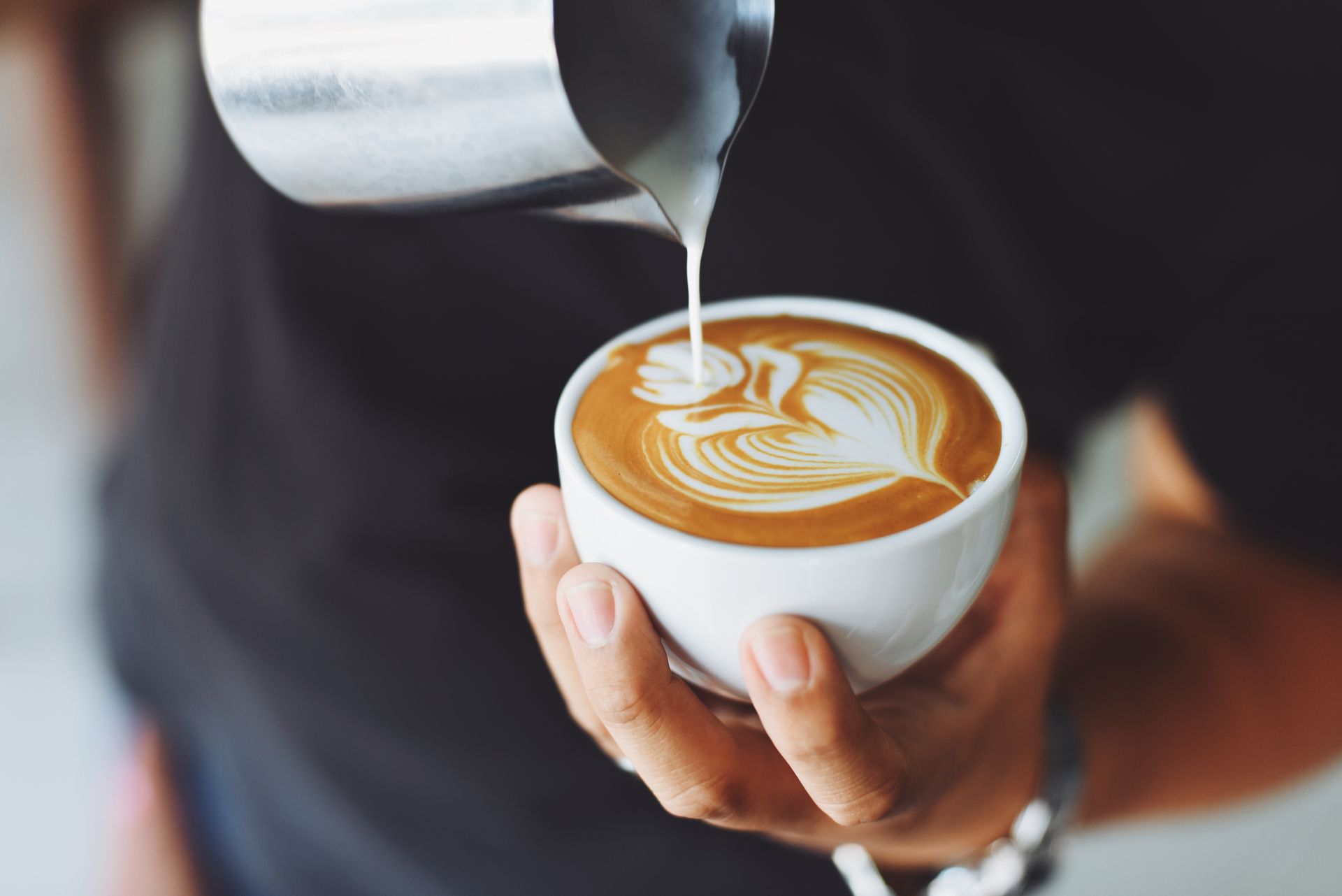 Barista pours milk into a latte with leaf latte art in a white cup.