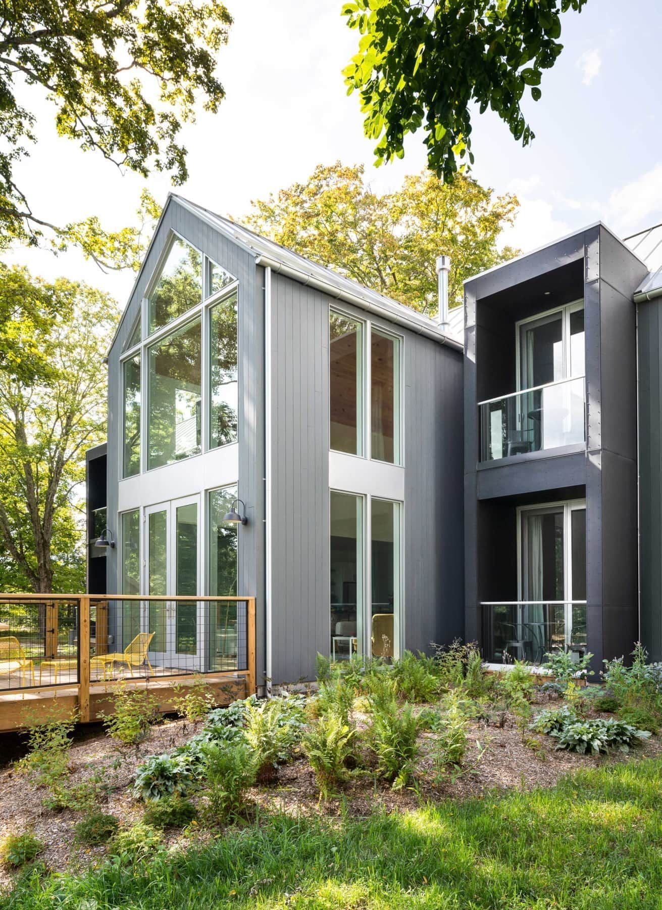 Modern glass-and-black house with tall windows, surrounded by trees and lush garden greenery