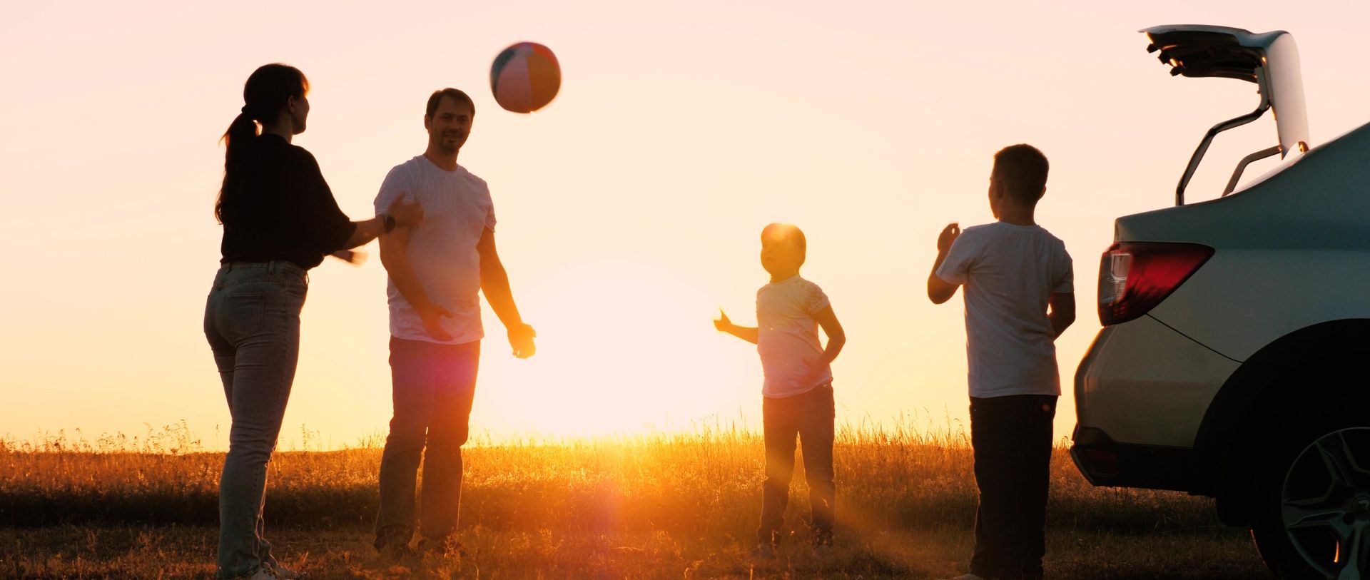Family playing with a ball outdoors near a car during sunset.