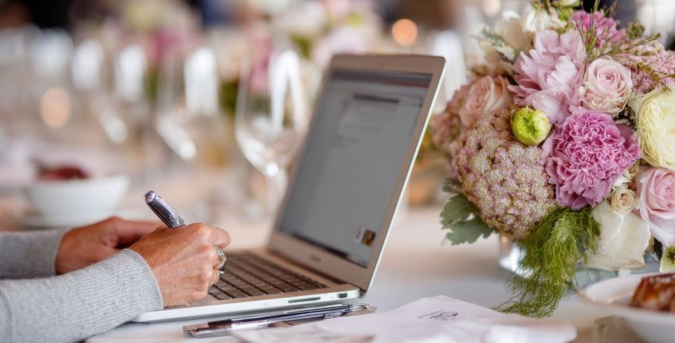Person using laptop, writing notes at a decorated table with flowers and wine glasses.