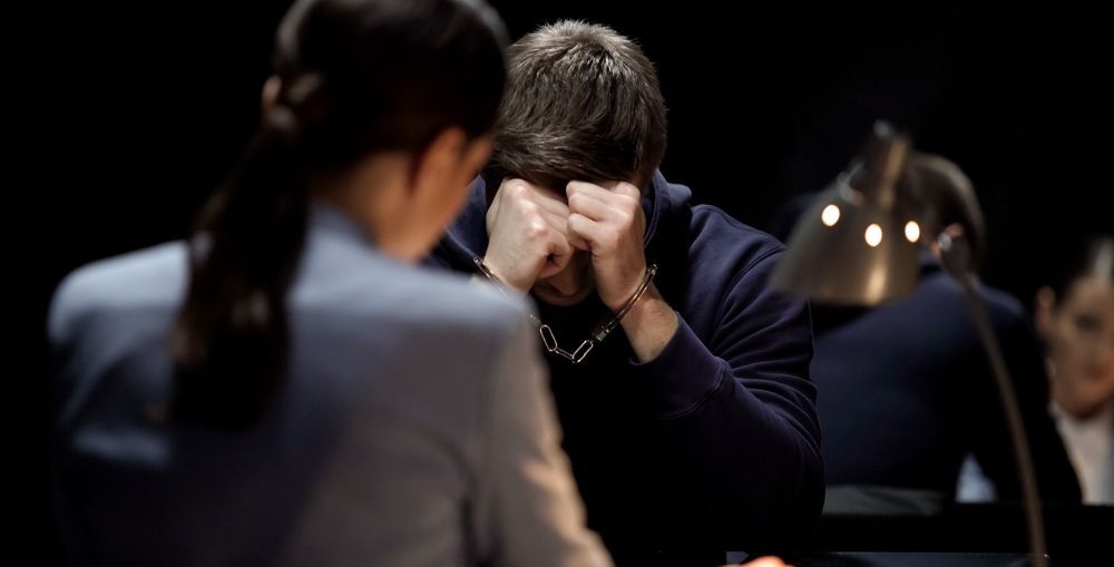 Man in handcuffs covering his face in an interrogation room; a person faces him.