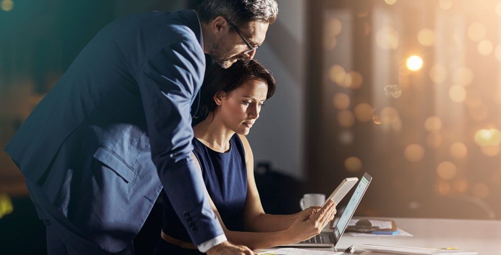A man in a suit and a woman in a dress looking at a laptop together at a desk.