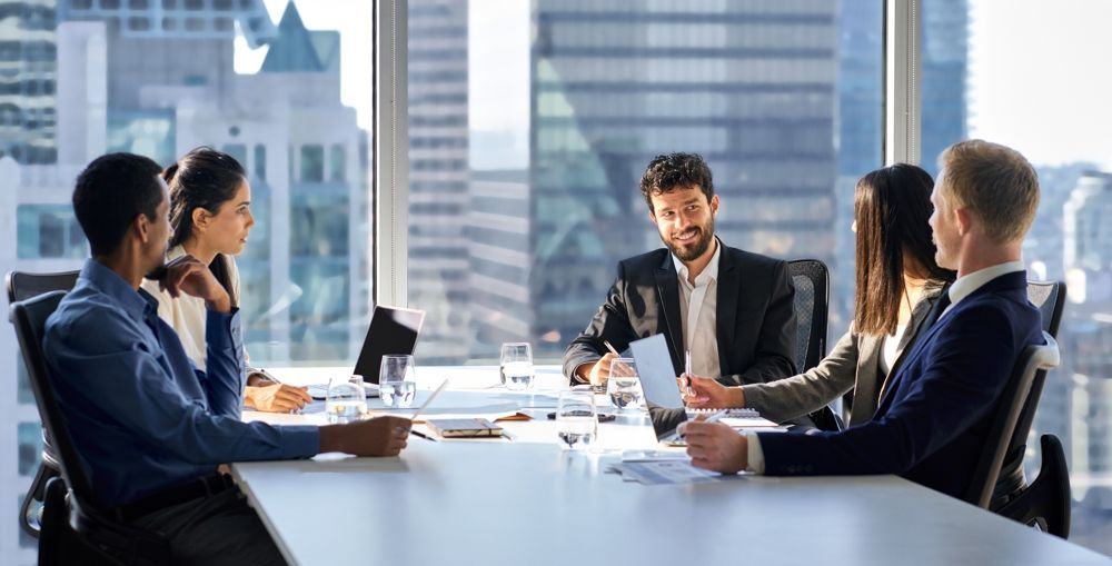 Businesspeople in a meeting at a table near a window, discussing documents and using tablets in an office.