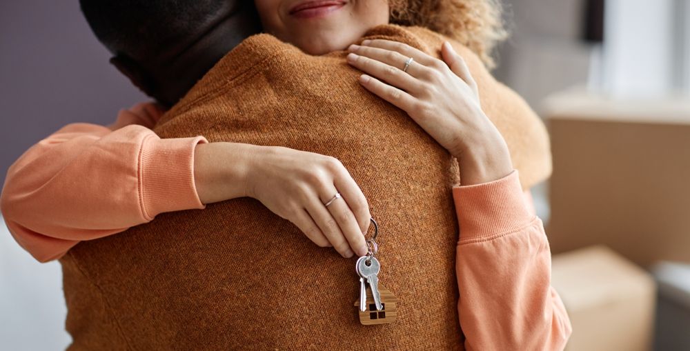 Woman hugging a person, holding keys; an orange sweater, brown sweater, and boxes are visible.