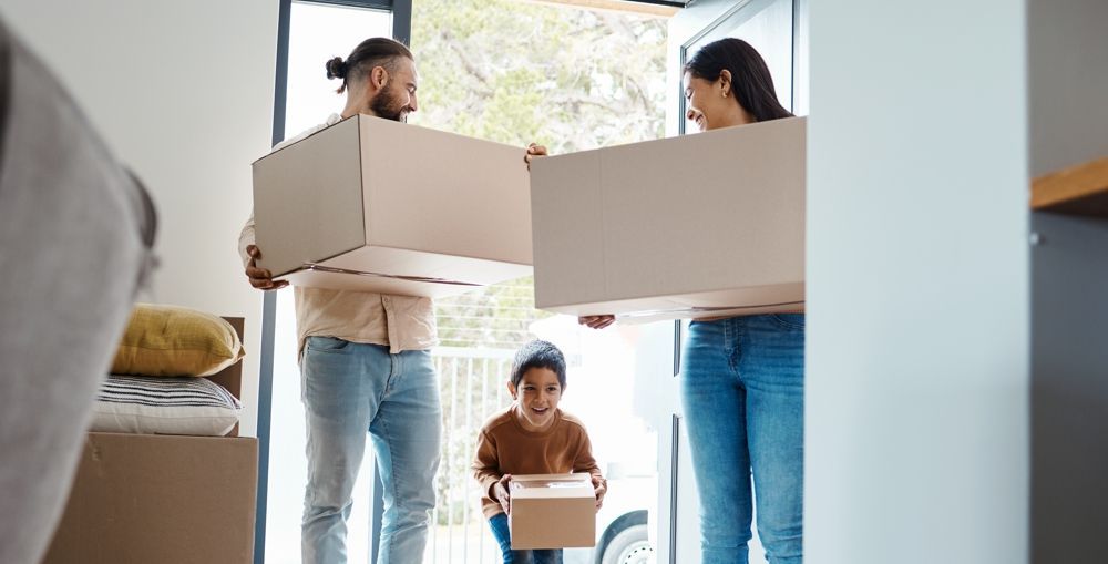 Family carrying boxes into a new home; child holds a small box; doorway with natural light.