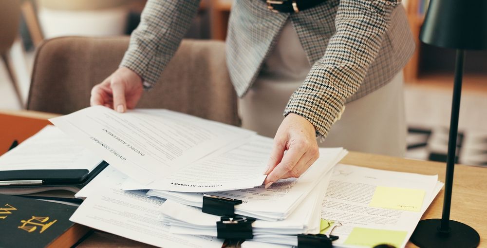 Person in blazer sorts through paperwork on a desk, in an office setting.