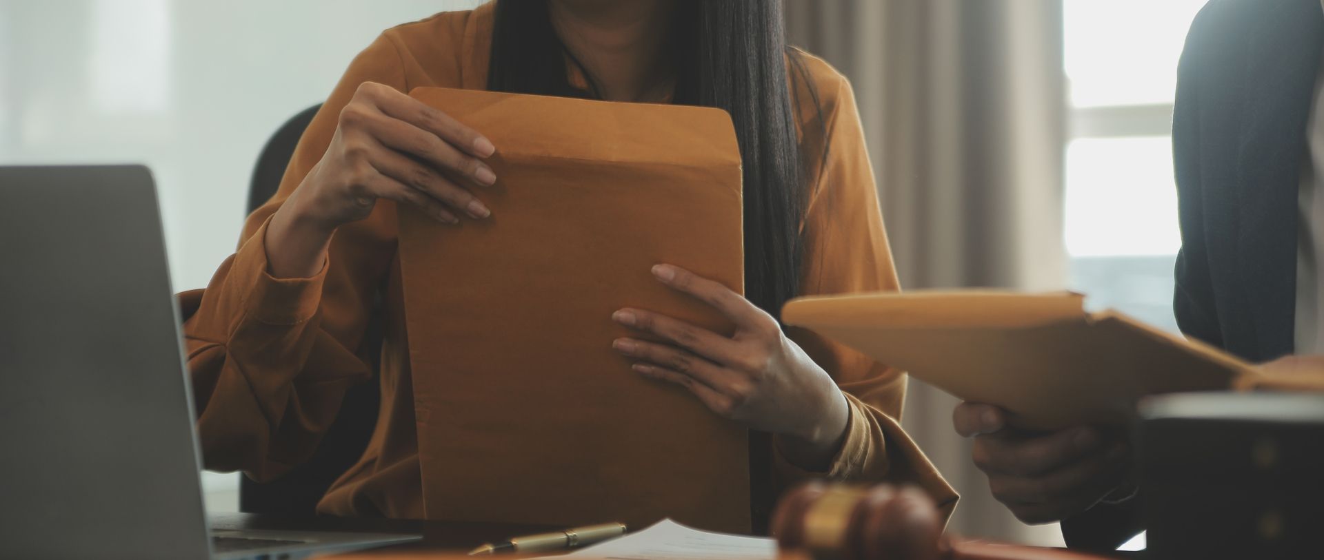 Woman in an orange shirt holding a large brown envelope, possibly in an office setting.