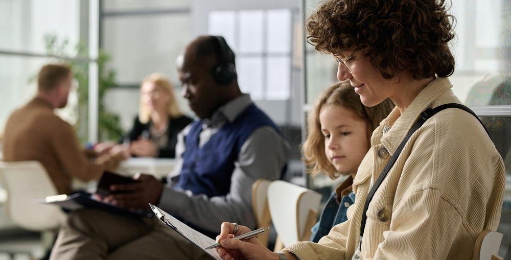 Woman with child filling out paperwork in a waiting room. Other people are in the background.