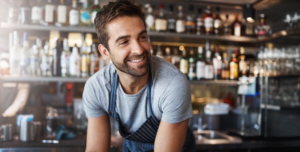 Bartender with a beard smiles, leaning on bar with liquor bottles in background.
