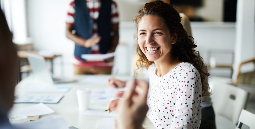 Woman smiling in a meeting with colleagues; light-colored shirt, sunny office.