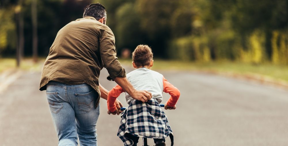 Father helping a child learn to ride a bike on a paved road.