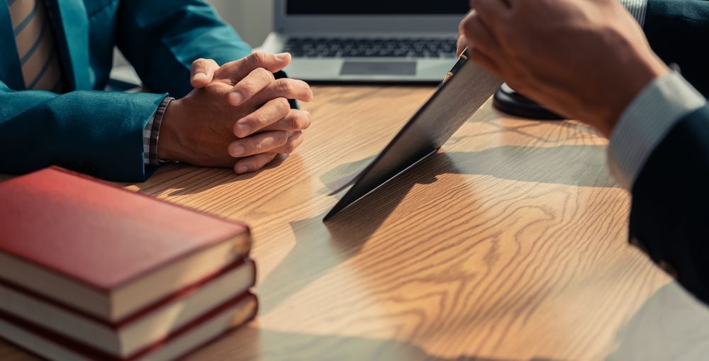 Two people in suits at a table, one presenting a file while the other has hands clasped. Books are on the table.
