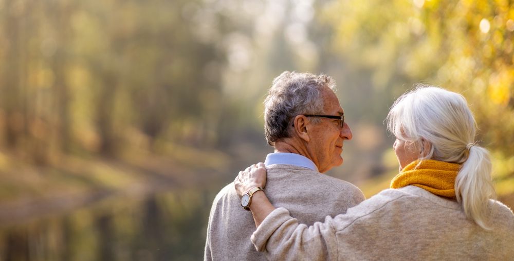 An elderly couple, backs to viewer, arm around each other, looking at a fall landscape.
