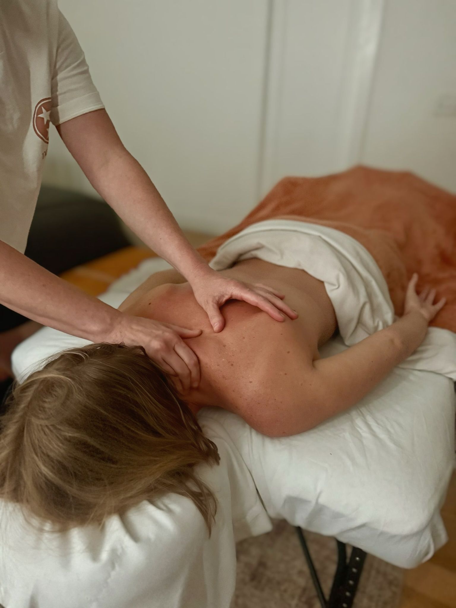 Person receiving a back massage on a table. Therapist's hands on the person's neck and back; white linens.