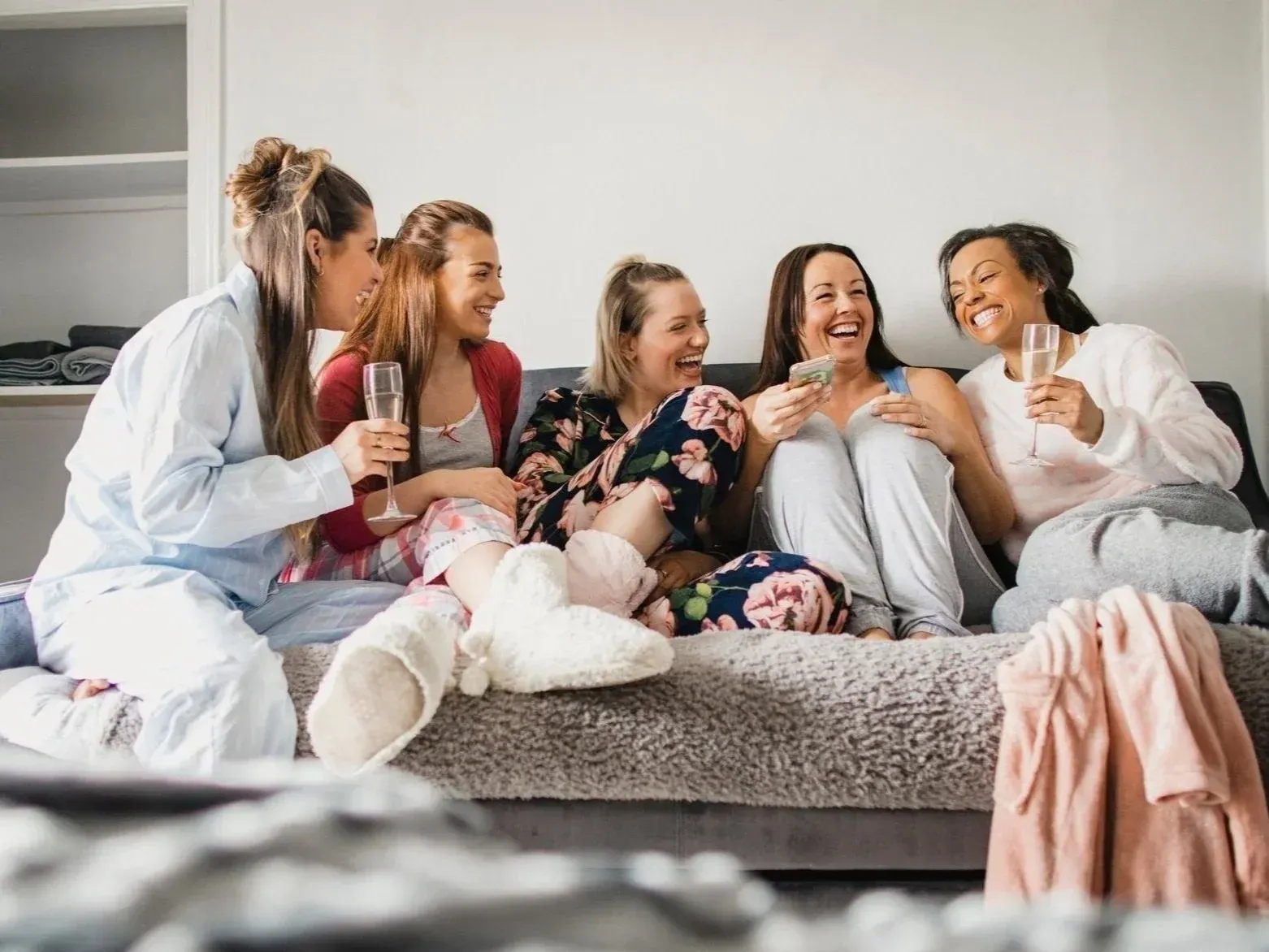 Five women in pajamas laughing, holding drinks, sitting on a sofa.