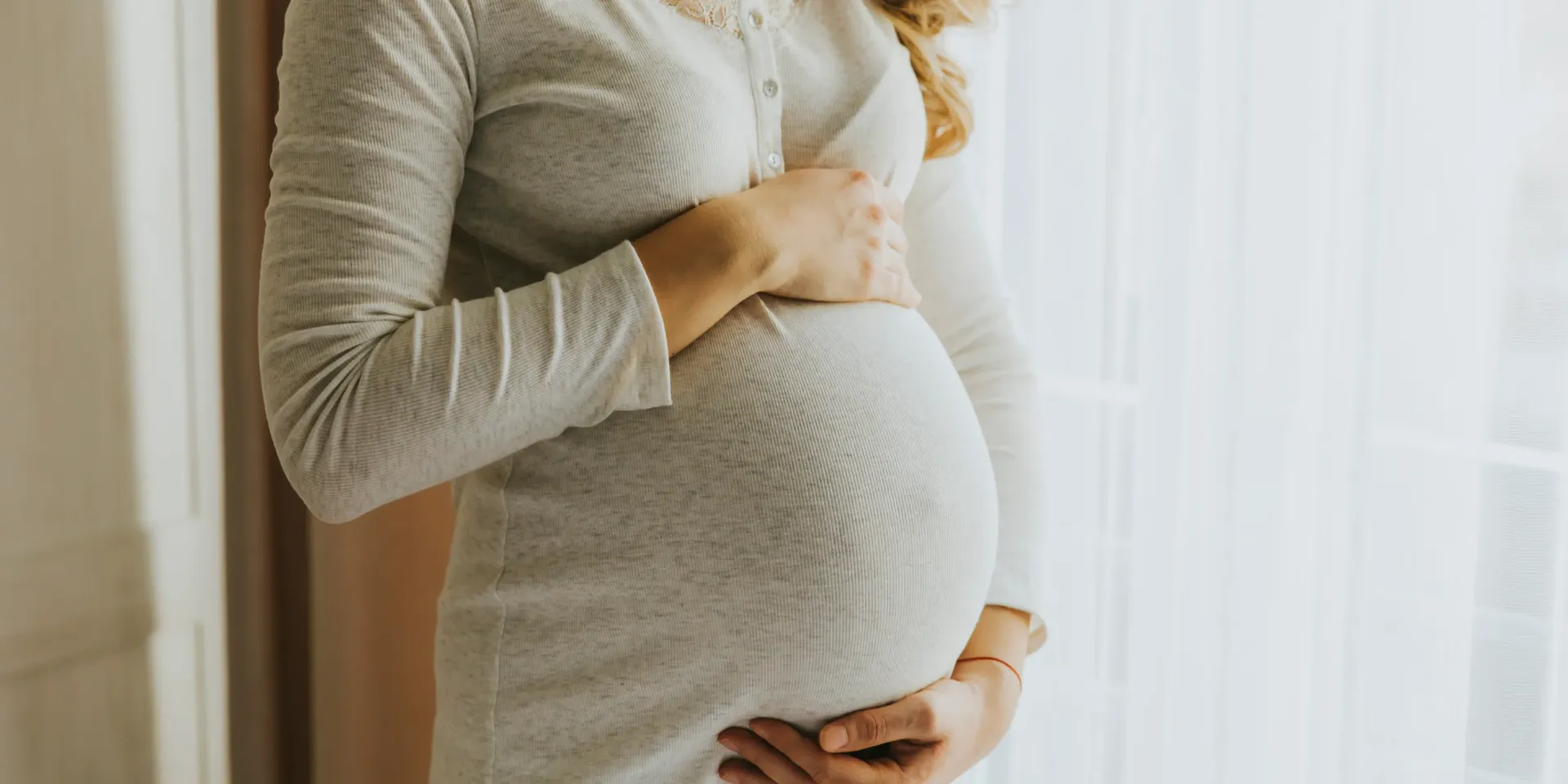 Pregnant woman in a light gray dress, holding her belly, near a window.
