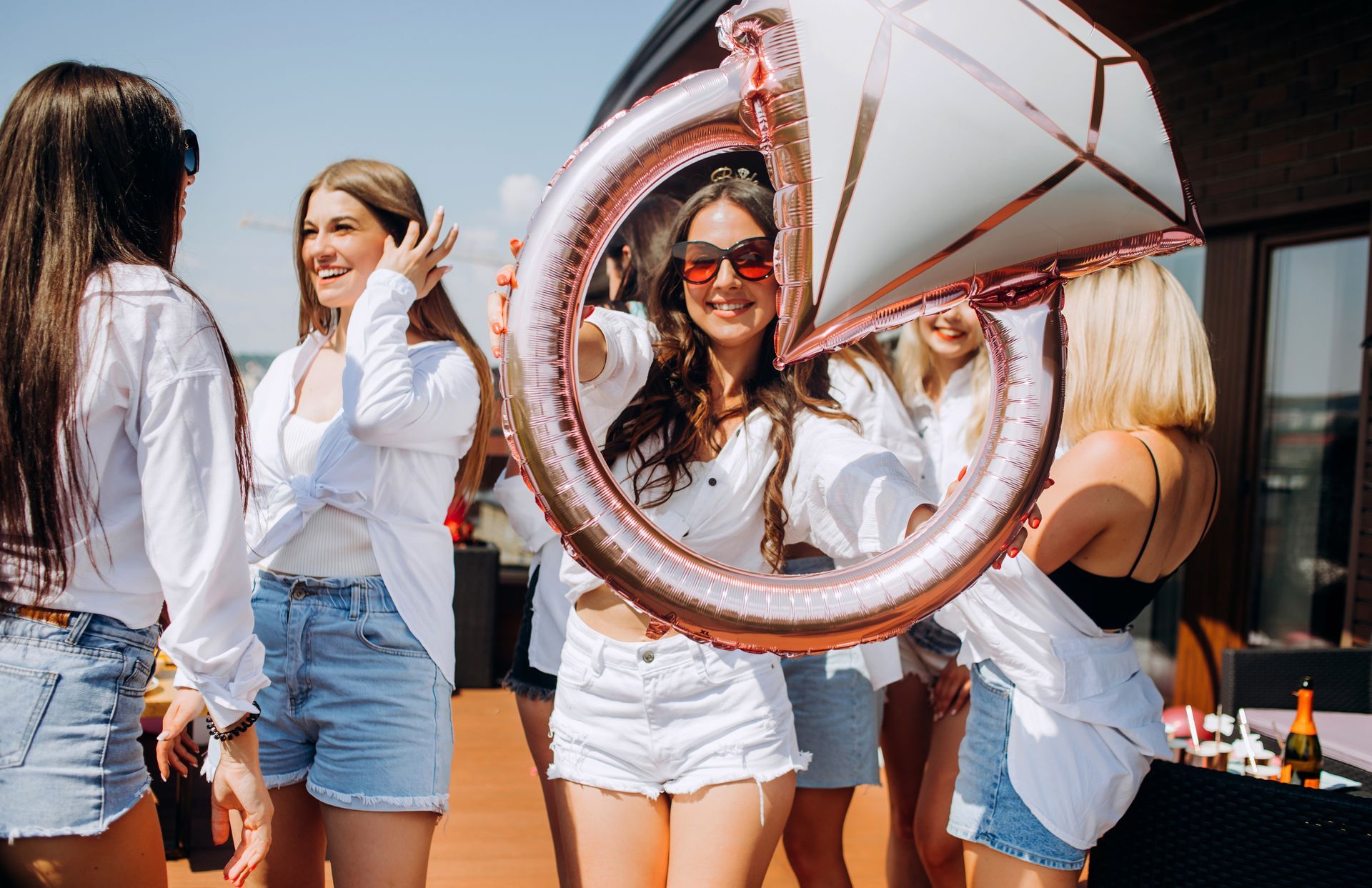 Women in white shirts and denim shorts at a rooftop bridal shower, holding a diamond ring balloon, smiling.