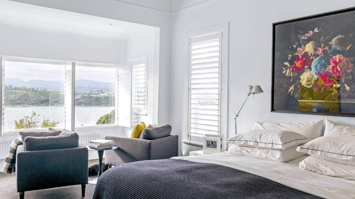 Bright modern bedroom with white walls, gray chairs, a bed, and a colorful floral painting above the headboard.