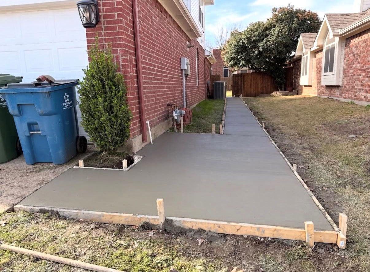Fresh concrete side yard walkway beside a brick house, with forms still in place and trash bins nearby