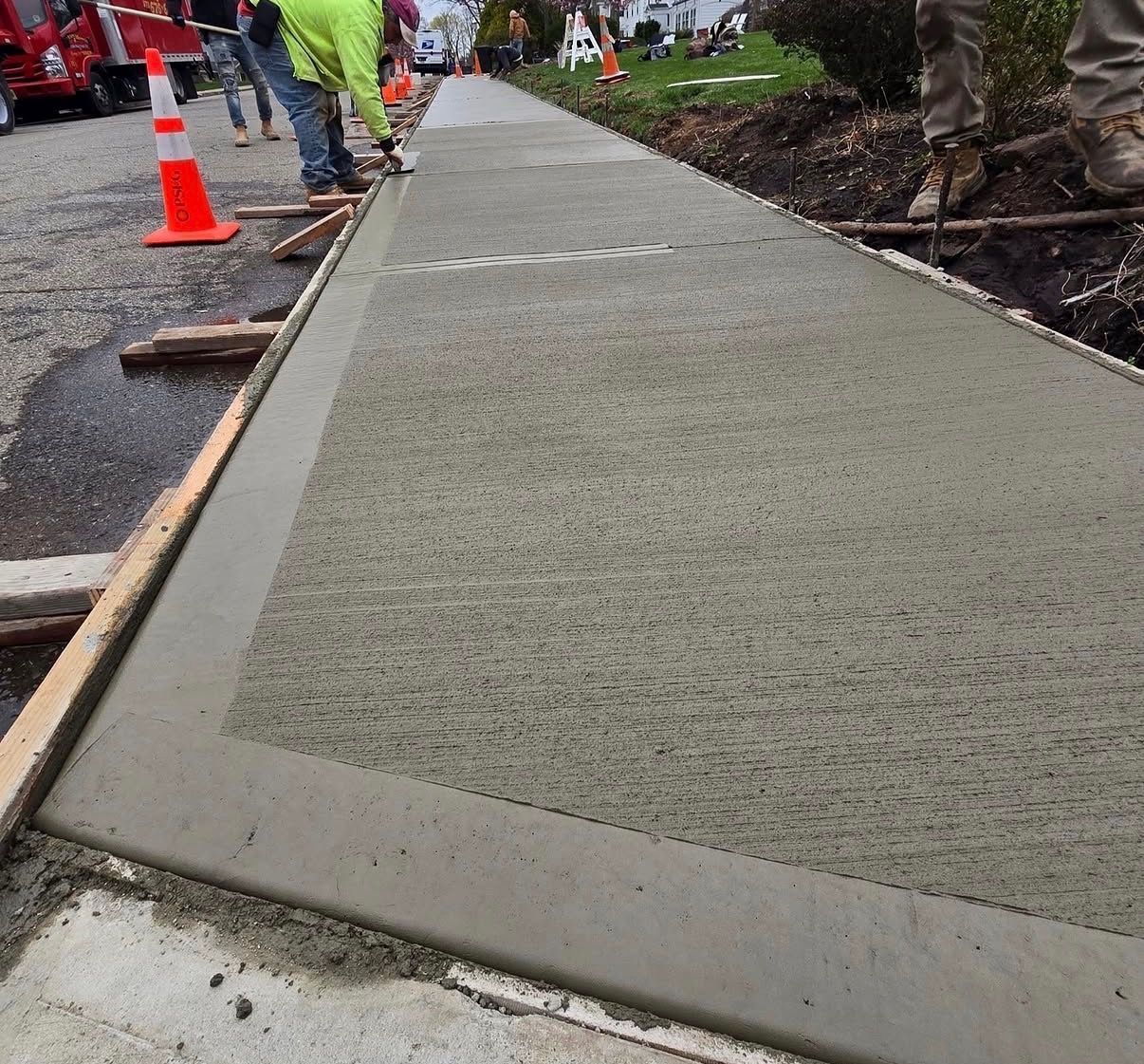 Freshly poured concrete sidewalk under construction with workers and orange safety cones nearby