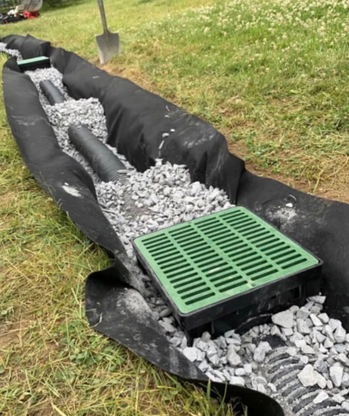Green storm drain grate in a black-lined gravel trench on grass, with a shovel nearby.
