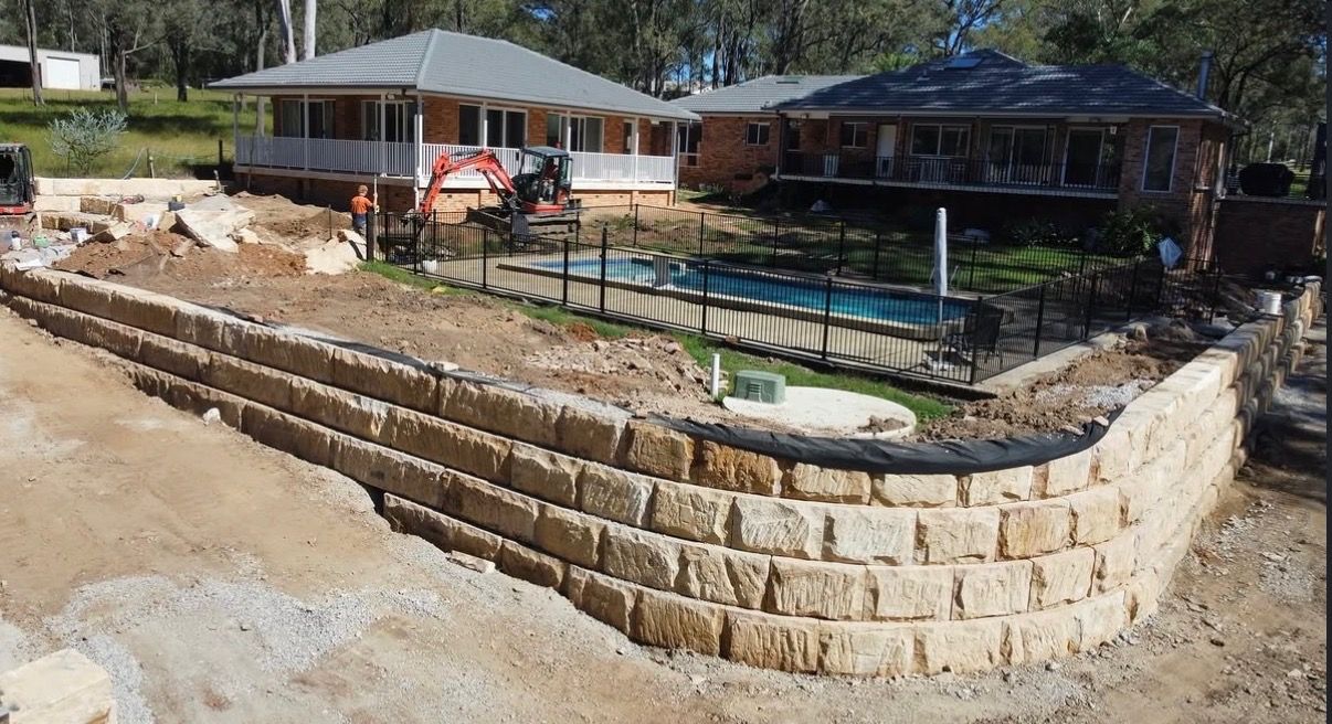 Retaining wall and landscaped pool area beside houses, with stone steps and an excavator nearby