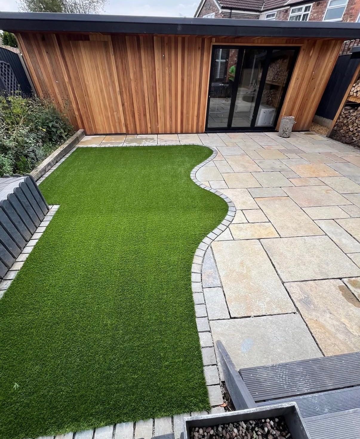Backyard patio with artificial grass, paver patio, and a wooden garden shed with glass doors