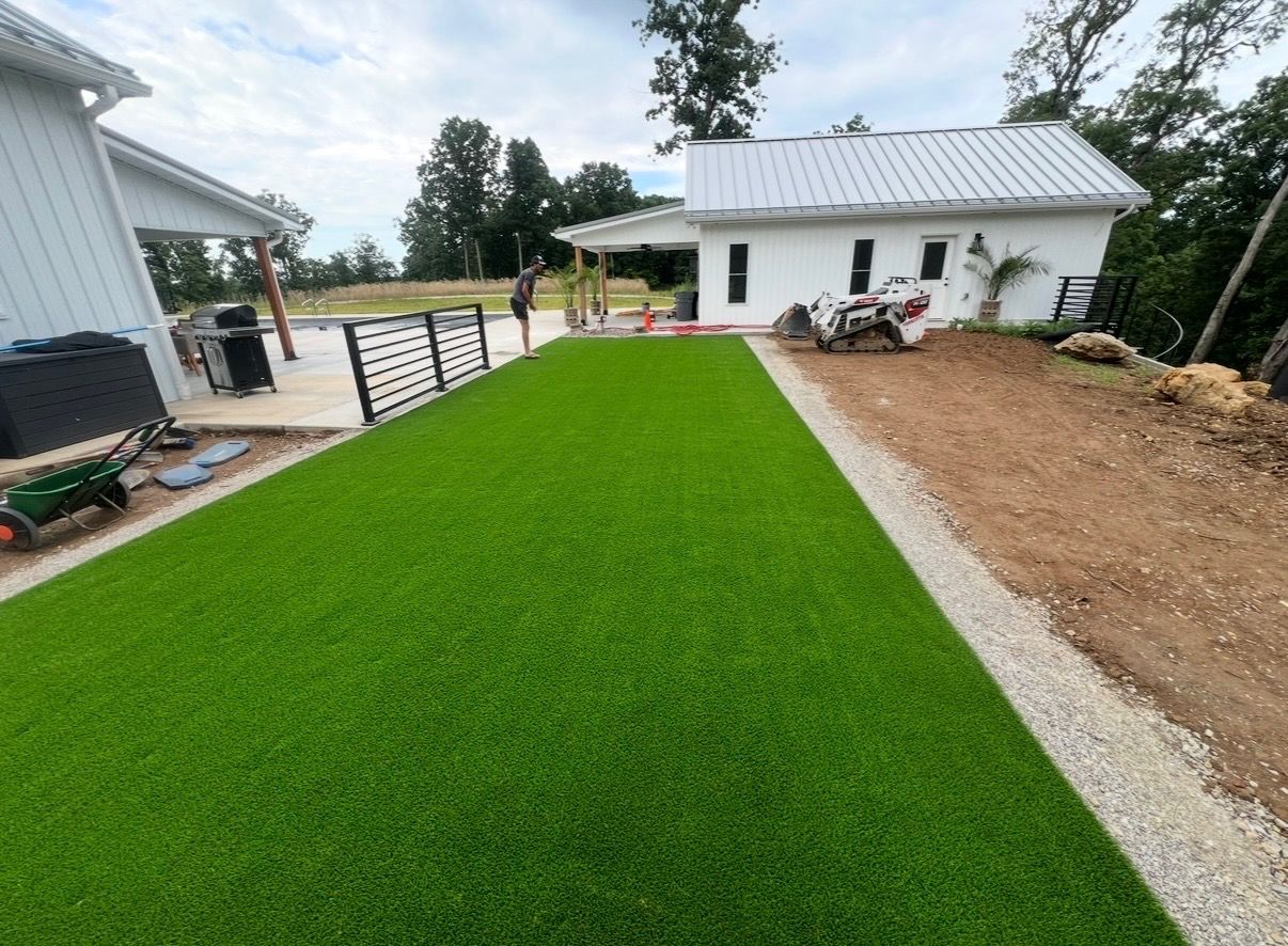 Bright green artificial turf beside a house, with a white building and gravel area in the background.
