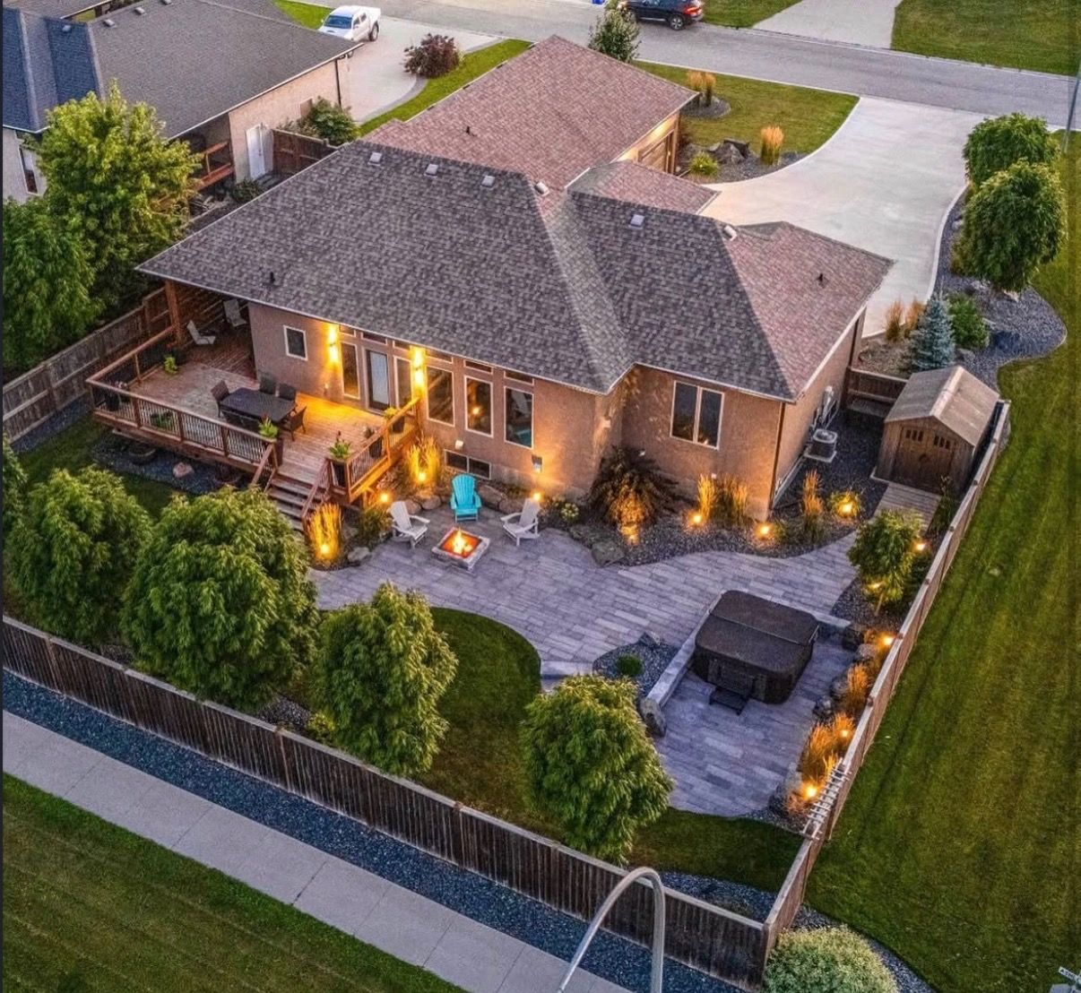 Aerial view of a suburban house with a lit patio, fenced yard, and driveway at dusk