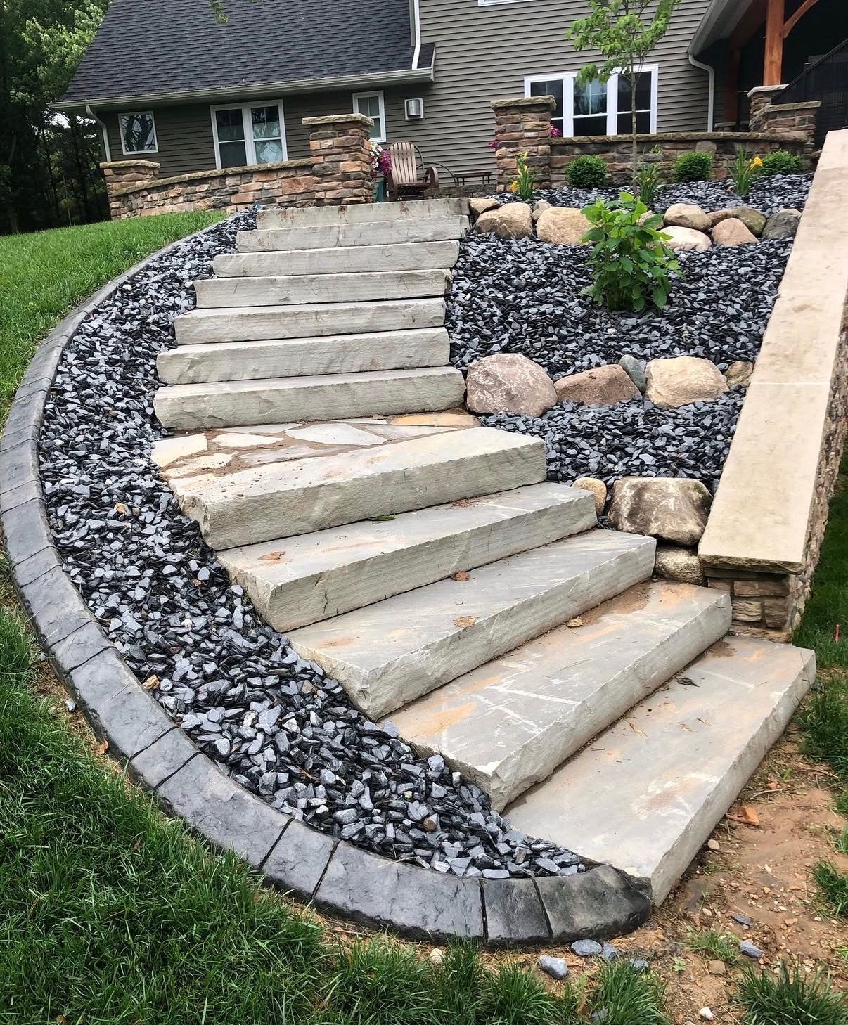 Curved stone steps leading to a house, bordered by black rocks and garden beds