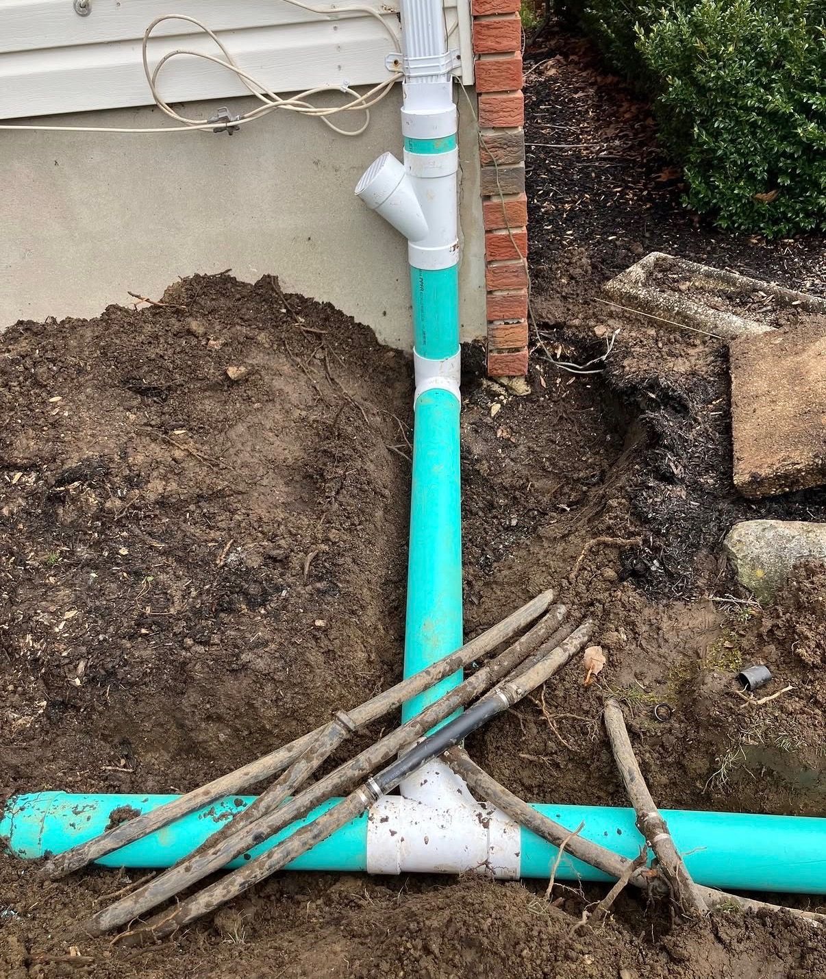 Drainage downspout with dirt piles and wooden stakes in a yard.