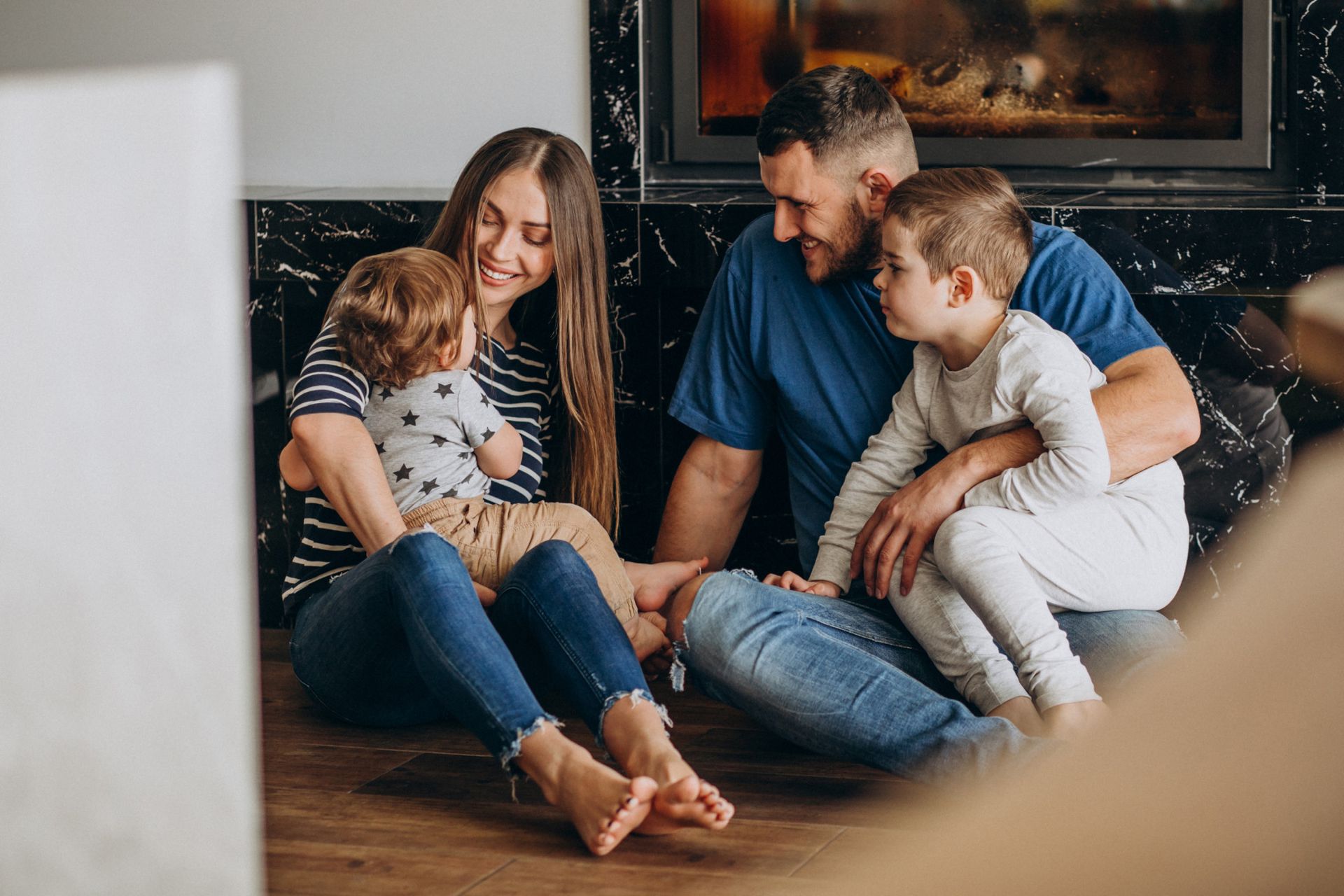 Family of four sitting on floor, smiling. Woman holds a baby; man sits with a child. Fireplace in background.