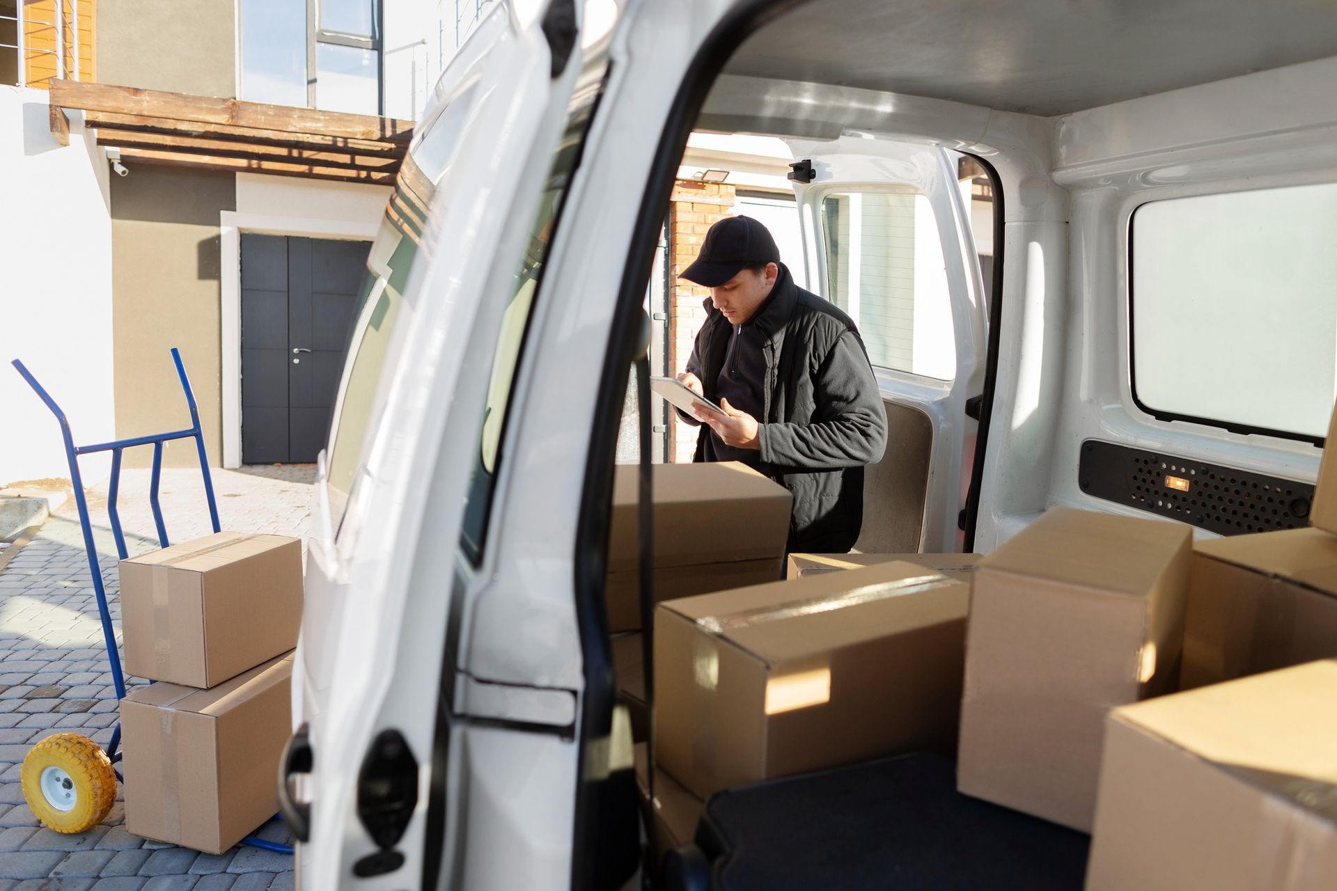 Delivery person in a van, checking a tablet amid cardboard boxes, next to a building.