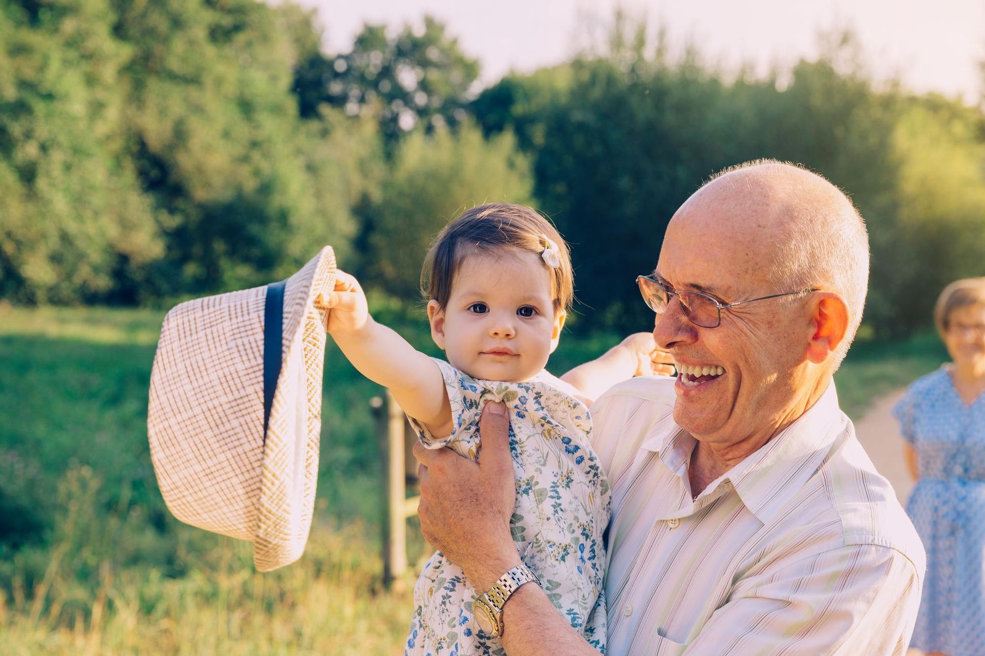 Smiling man holding a baby waving a hat outdoors; woman walking in background.