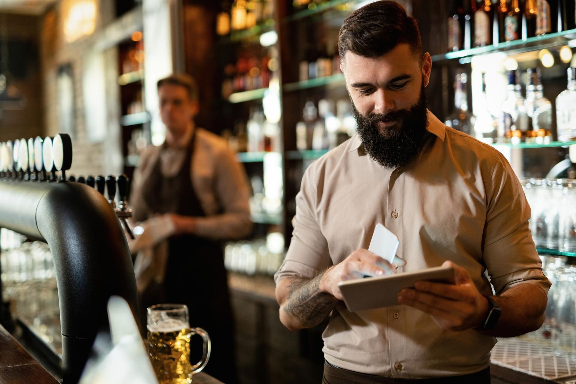 Bartender with a beard using a tablet behind a bar. Another bartender in the background.