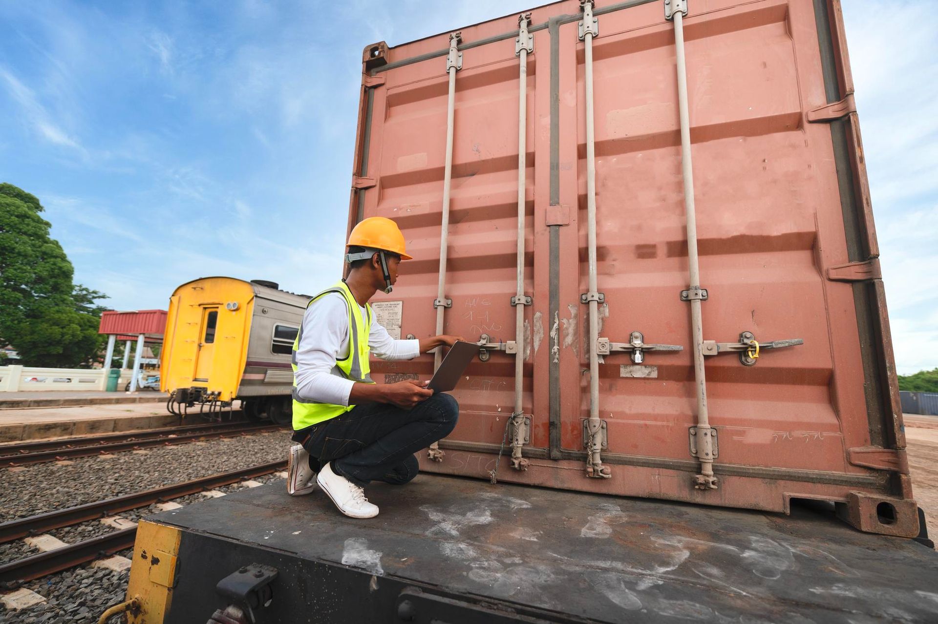 Man in safety vest and helmet inspects cargo container on train with a laptop.