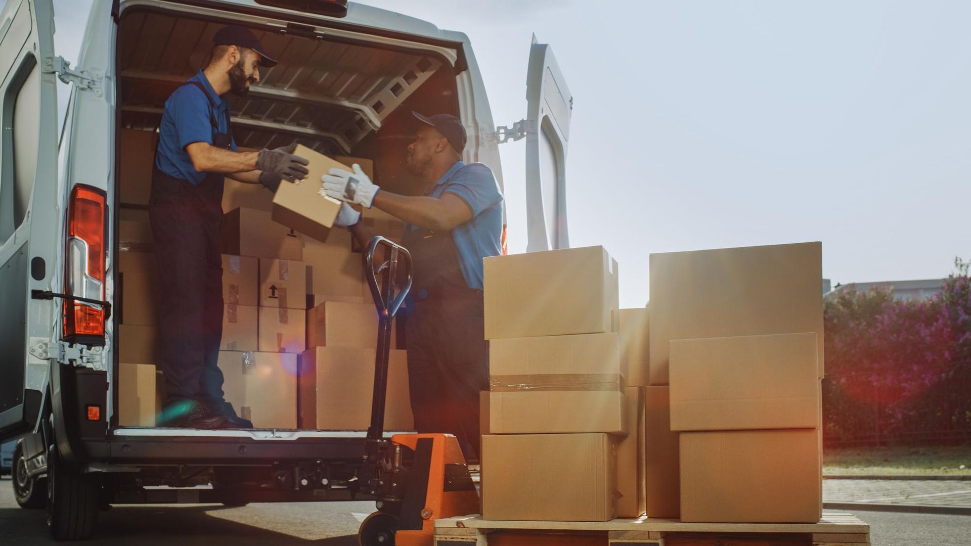 Two workers loading cardboard boxes into a delivery van using a pallet jack outdoors.