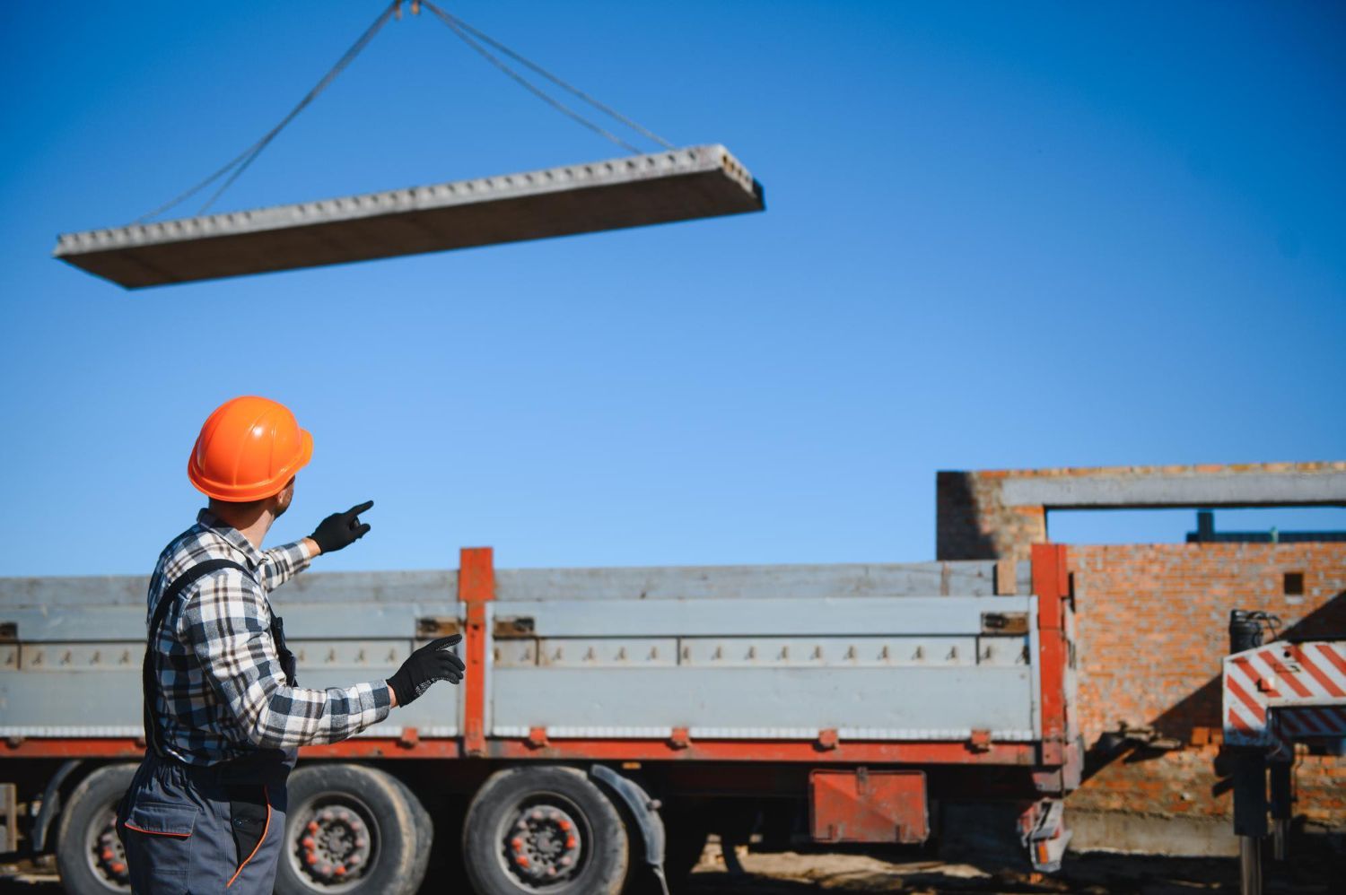 Construction worker in hard hat directs a concrete slab being lifted by a crane over a truck bed against a blue sky.