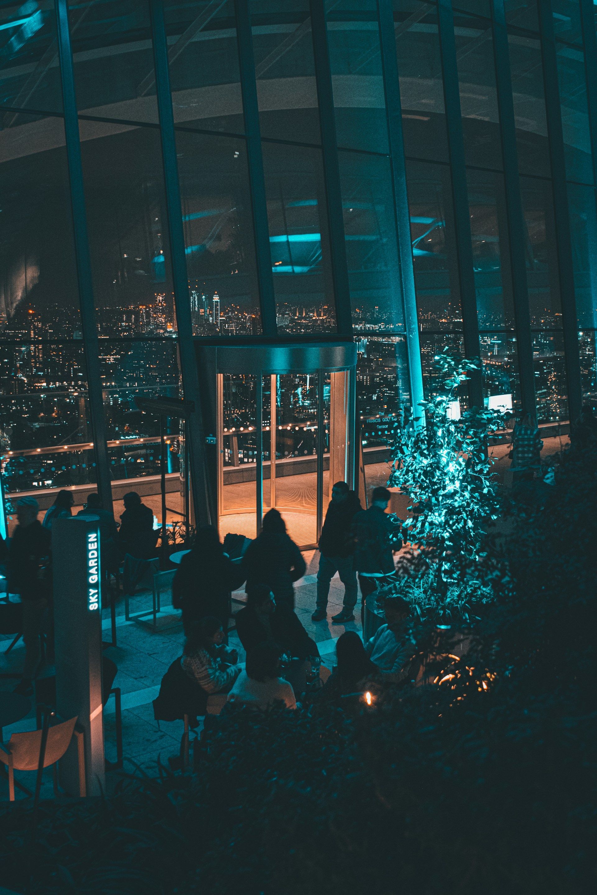 People in a glass building at night, city lights visible. Blue ambient lighting.