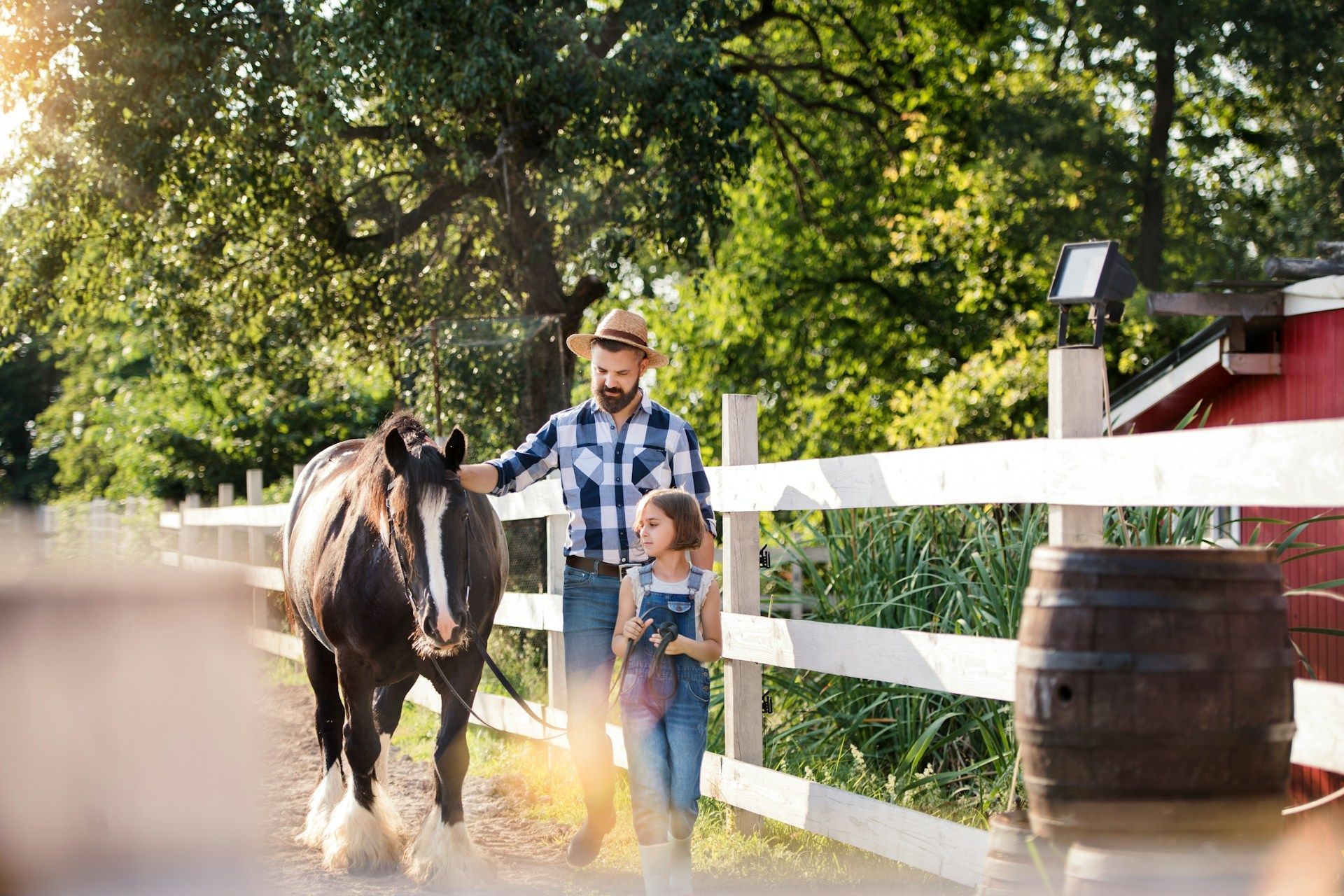 Man and child walking a horse on a farm; a white fence, red building, and wooden barrel are also present.