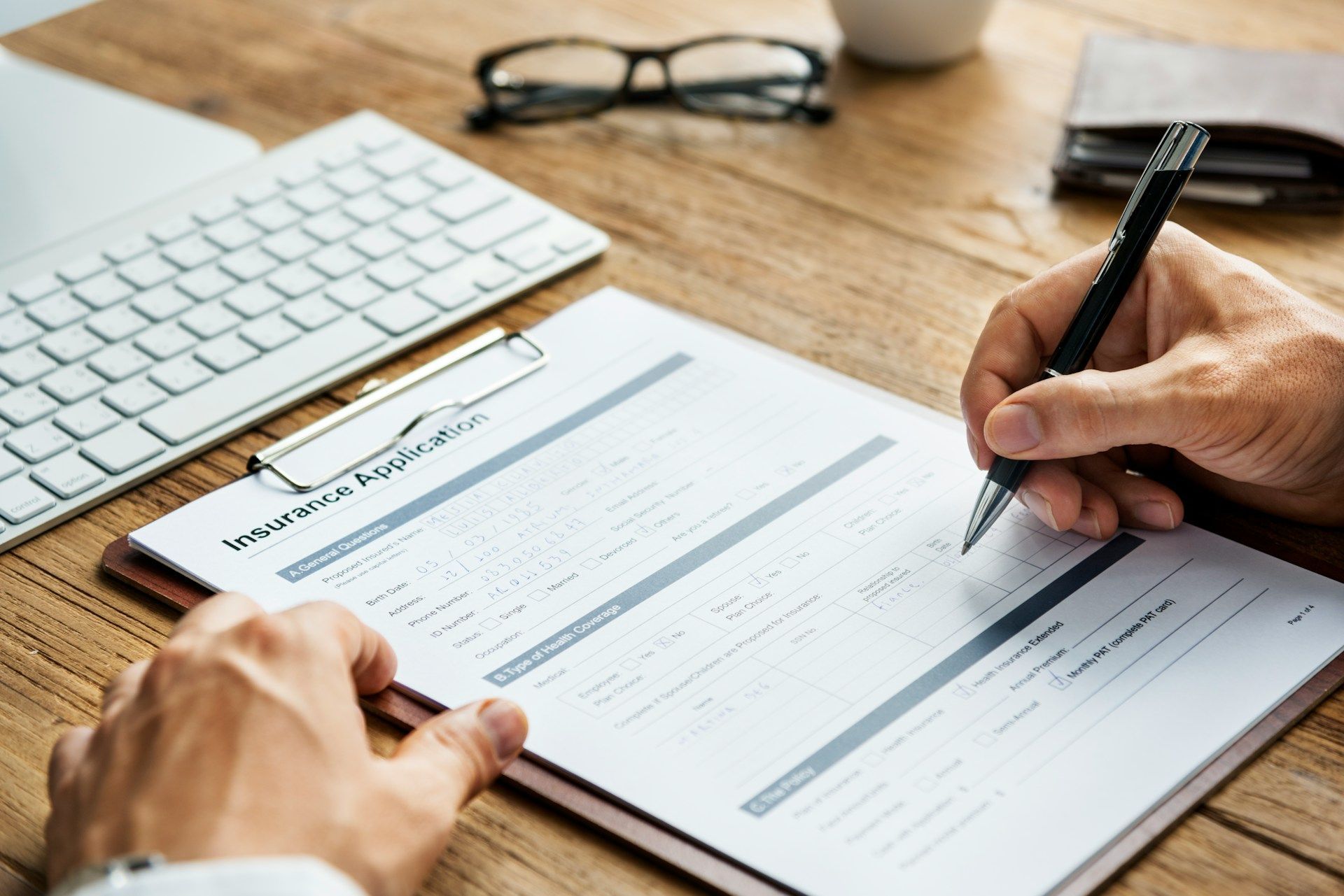 Person filling out a form with a pen on a wooden desk with keyboard and glasses.