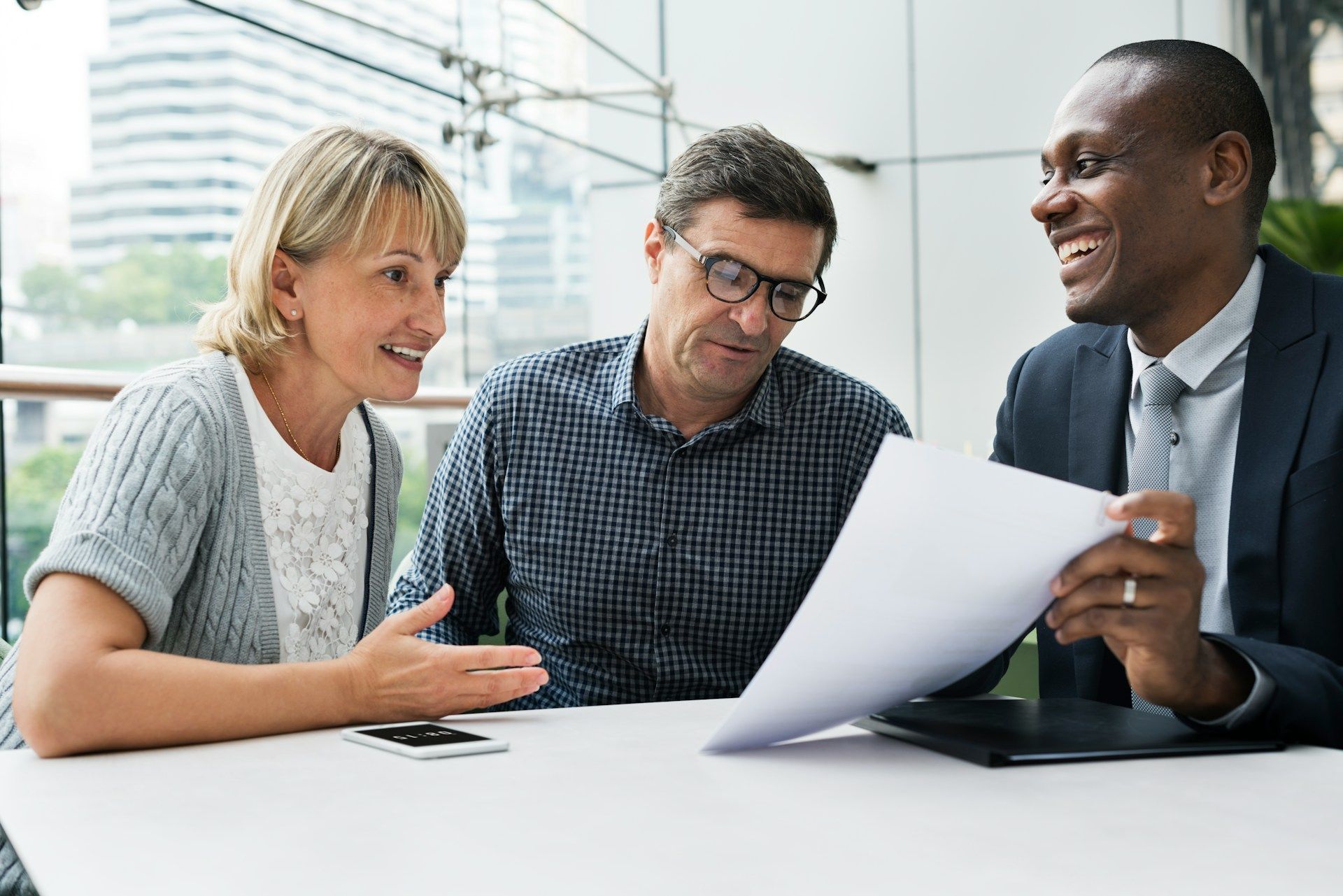 Three people review paperwork at a table; one smiles, pointing.