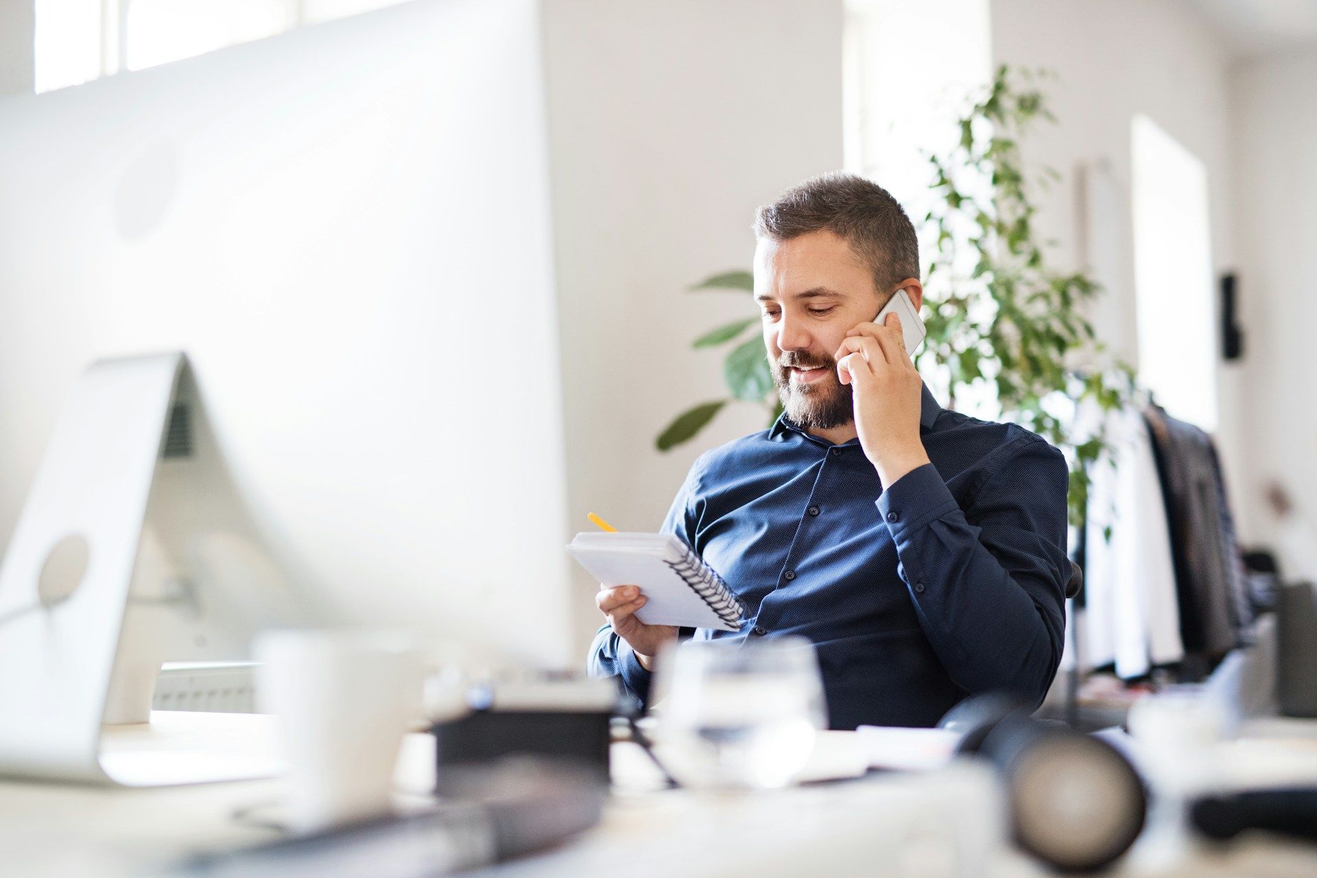 Man in blue shirt on phone, taking notes at a desk in an office setting.
