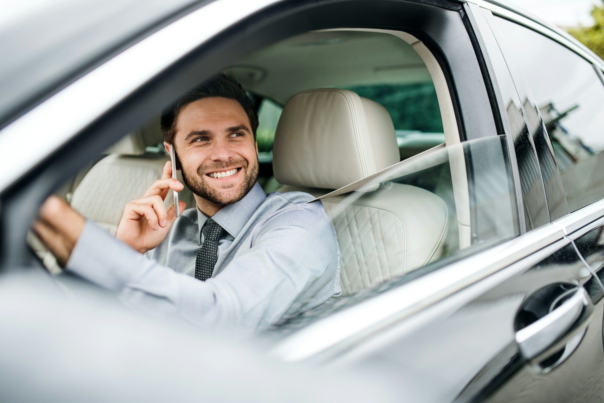 Man in a car smiling while talking on the phone. Grey car interior visible.