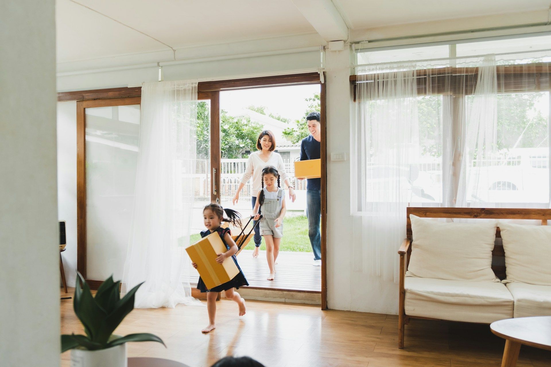Family entering a home with boxes. A girl runs ahead, smiling. Woman and child follow. Man stands in doorway.