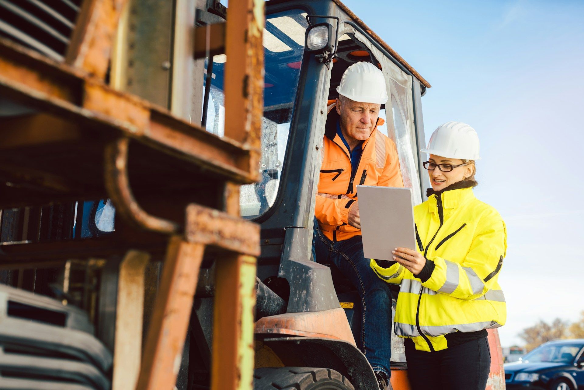 Two people in safety vests and hard hats reviewing a tablet near a forklift.